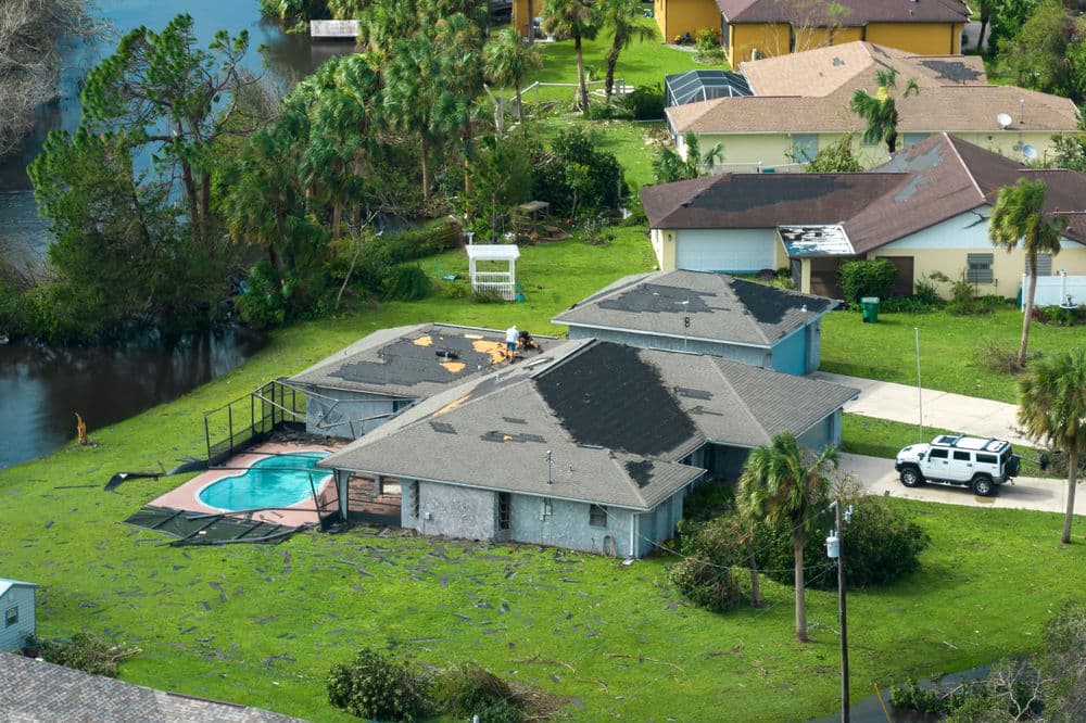 Aerial view of hurricane-damaged homes with roofs under repair, near a pool and water.