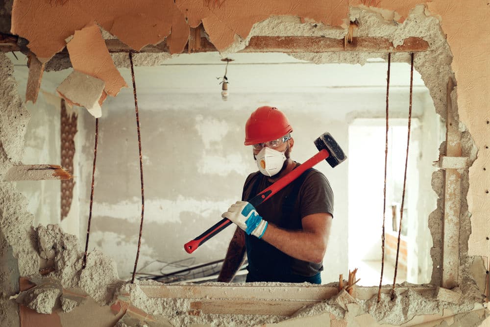 Construction worker in a hard hat and mask holding a sledgehammer, standing in a demolition site.
